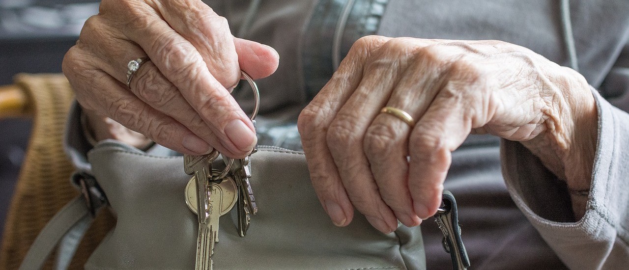 The picture shows the hands of an old lady sitting on a chair. She is wearing a diamond ring on her middle finger on her right hand and a gold ring on her ring finder on her left hand. She wears a grey top and puts several keys on a key ring in a grey handbag on her lap.