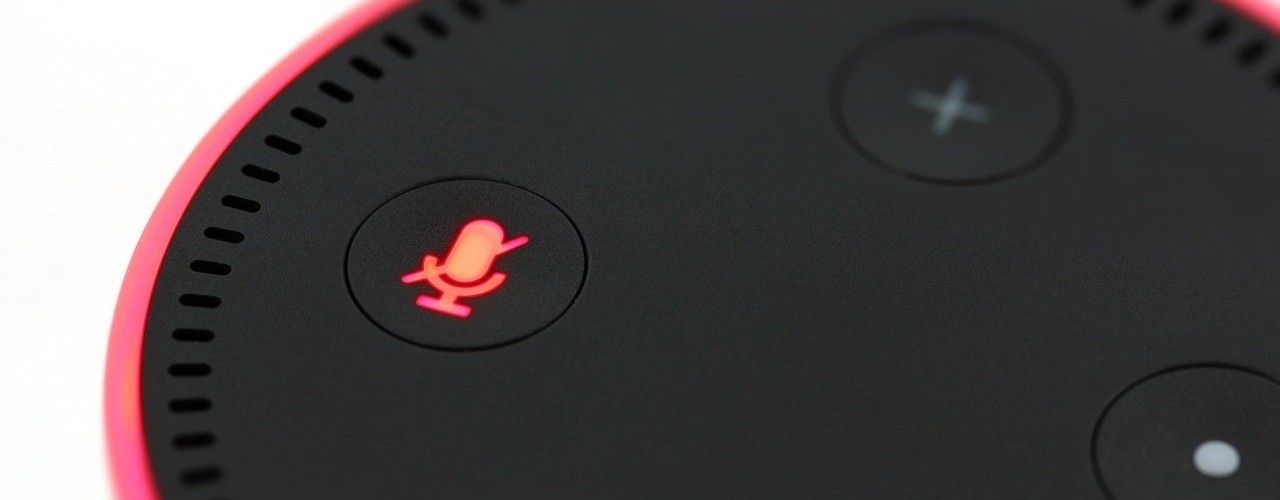 Auf dem Bild ist ein Amazon Echo Dot mit roter Beleuchtung zu sehen. Es handelt sich dabei um einen kreisförmigen, flachen Lautsprecher, der auf Sprachbefehle reagiert. Die Taste, mit der man das Mikrophon auf stumm stellen kann, leuchtet rot. 