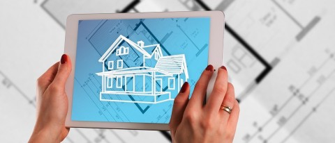 The picture shows the hands of a woman. She is wearing red nail polish and a wide ring on her right ring finger. She is holding a white tablet in her hands on which a sketch of a house can be seen. The screen background is blue and shows the ground plan of a building. The ground plan is also on paper on a table under the tablet. The sketch of the house looks like a projection of the finished building over of the building's ground plan.