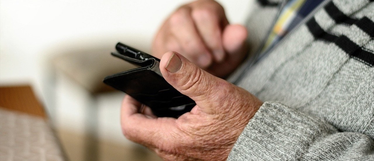 The picture shows the hands of an old man. He is wearing a grey jumper with two black stripes across his chest and is holding a mobile phone in his hands. He seems to be looking at it. Old-fashioned furniture can be seen in the background.