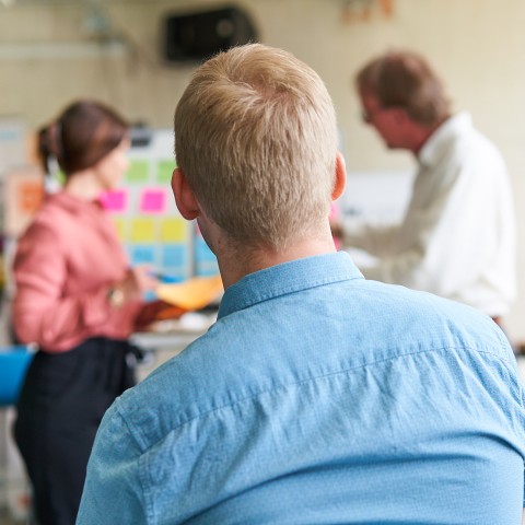 Eine Frau und ein Mann arbeiten mit Post-Its an einem Whiteboard, eoin Mann sitzt davor