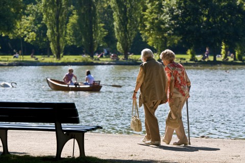 Zwei Seniorinnen spazieren am See, eine Bank ist im Vordergrund zu sehen, auf dem See ein Ruderboot mit zwei jungen Menschen