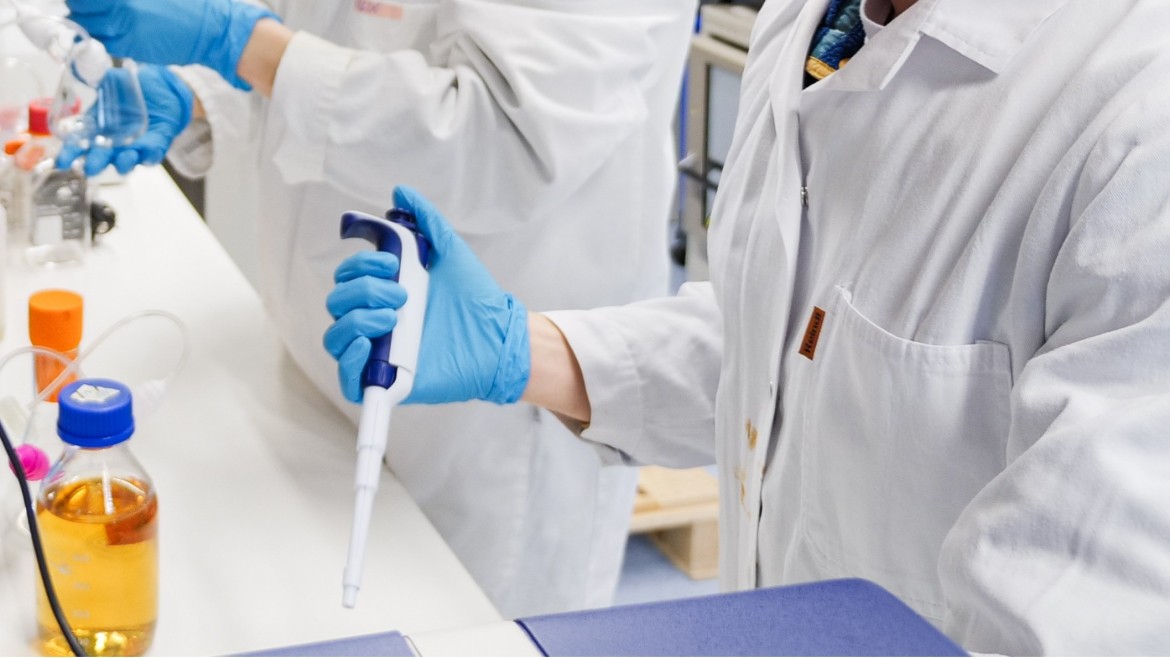 Zu sehen sind zwei Personen, die molekularbiologische Arbeiten an einem Laborarbeitsplatz verrichten. Eine Person hält eine Pipette in der Hand, die andere Person einen Erlenmeyerkolben.