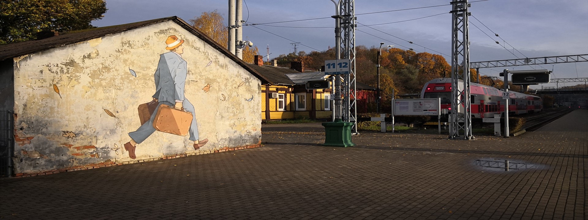 Kleiner Bahnhof im Herbst, links im Vordergrund ein Gebäude mit Gemälde - ein Mann im Anzung und Hut trägt zwei Koffer und läuft Richtung Gleis. Im Hintergrund steht ein roter Zug, daneben ein kleines gelbes Gebäude und Bäume. 