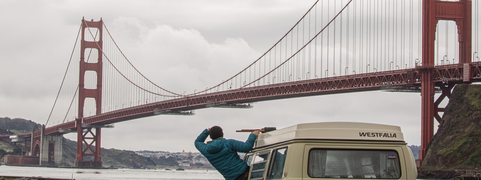 Bewölkter Himmel, im Hintergrund die Golden Gate Bridge in San Francisco. Am Flußufer steht ein grau-grüner Transporter, aus dem Fenster (Fahrersitz) lehnt sich ein Student in blauer Jacke und schaut Rocjtung der Brücke. 