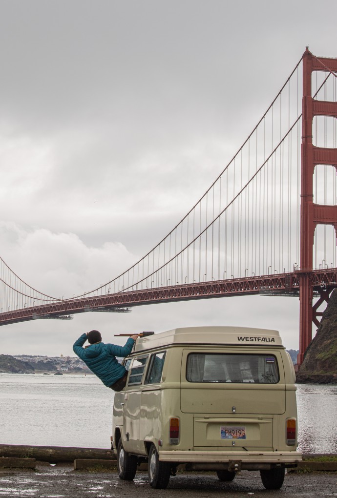 Bewölkter Himmel, im Hintergrund die Golden Gate Bridge in San Francisco. Am Flußufer steht ein grau-grüner Transporter, aus dem Fenster (Fahrersitz) lehnt sich ein Student in blauer Jacke und schaut Rocjtung der Brücke. 
