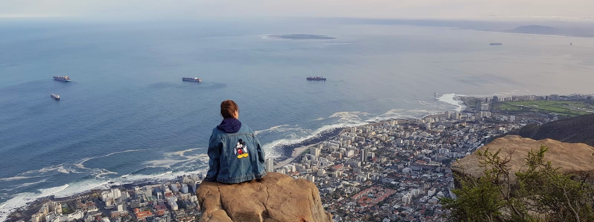 Eine Studentin sitzt auf einem Felsen, sie hat eine Jeansjacke mit Micky Mouse auf dem Rücken an. Sie blickt von oben auf Kapstadt. Die Stadt liegt am Ozean, den man im Hintergrund sieht. Man sieht auch mehrere Containerschiffe. Der Himmel ist bewölkt. 