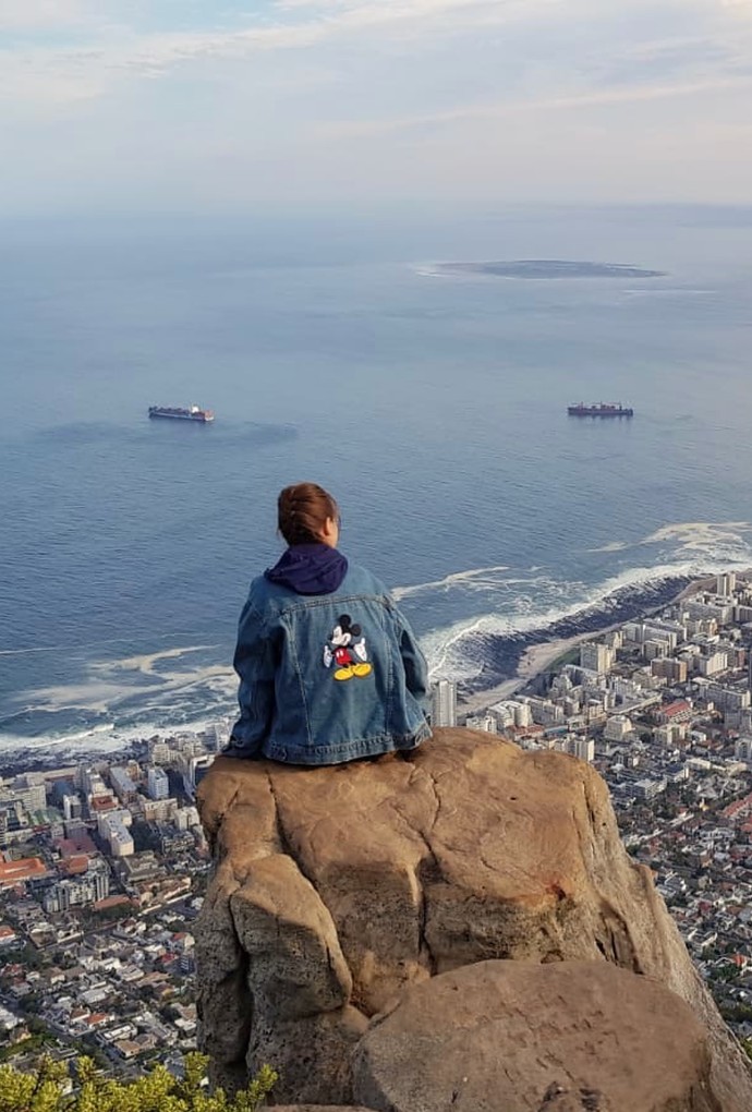Eine Studentin sitzt auf einem Felsen, sie hat eine Jeansjacke mit Micky Mouse auf dem Rücken an. Sie blickt von oben auf Kapstadt. Die Stadt liegt am Ozean, den man im Hintergrund sieht. Man sieht auch mehrere Containerschiffe. Der Himmel ist bewölkt. 