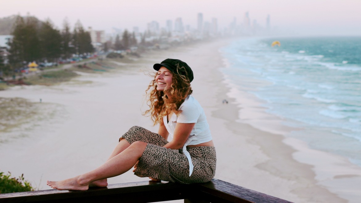 Eine lächelnde Studentin mit langen lockingen Haaren sitzt auf einem Balken. Sie hat ein schwarzes Cappy, weißes T-Shirt und Hose mit Leopardenmuster an. Im Hintergrund ein Sandstrand und Ozean sowie eine Großstadt mit Wolkenkratzern. 