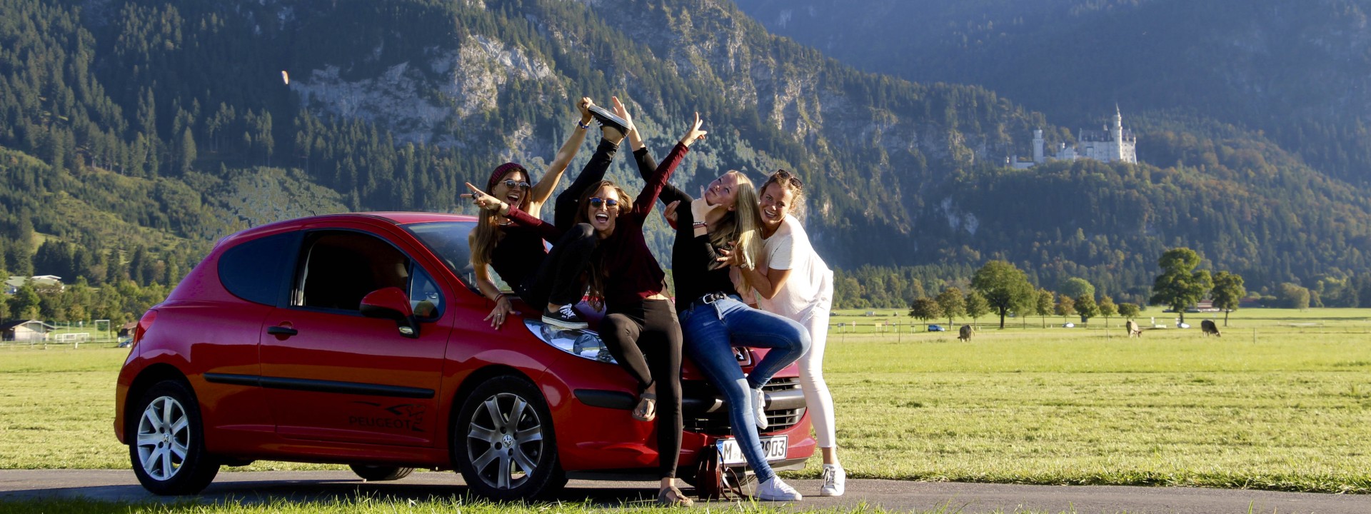 4 female students posing on the mask of a red small car, in the background a meadow, mountains and Neuschwanstein Castle
