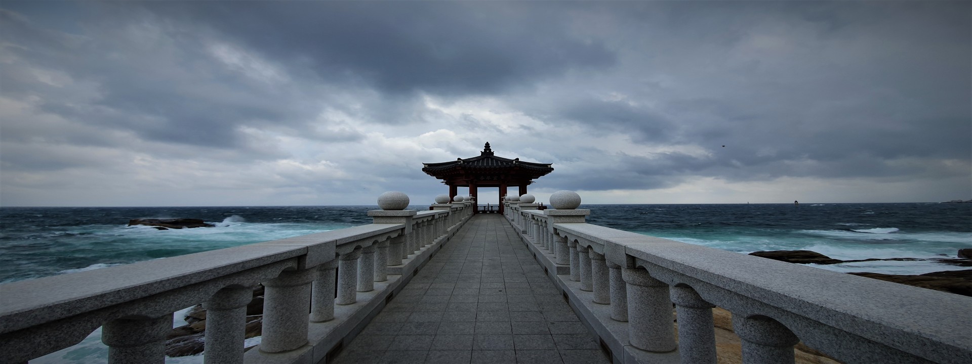 Im Vordergrund, mittig im Bild, sieht man eine Hafenmole aus Marmor. Am Ender der Mole steht ein typischer chinesicher Pavillon. Im Hintergrund aufgewühltes Meer und stark bewölkter Himmel. 