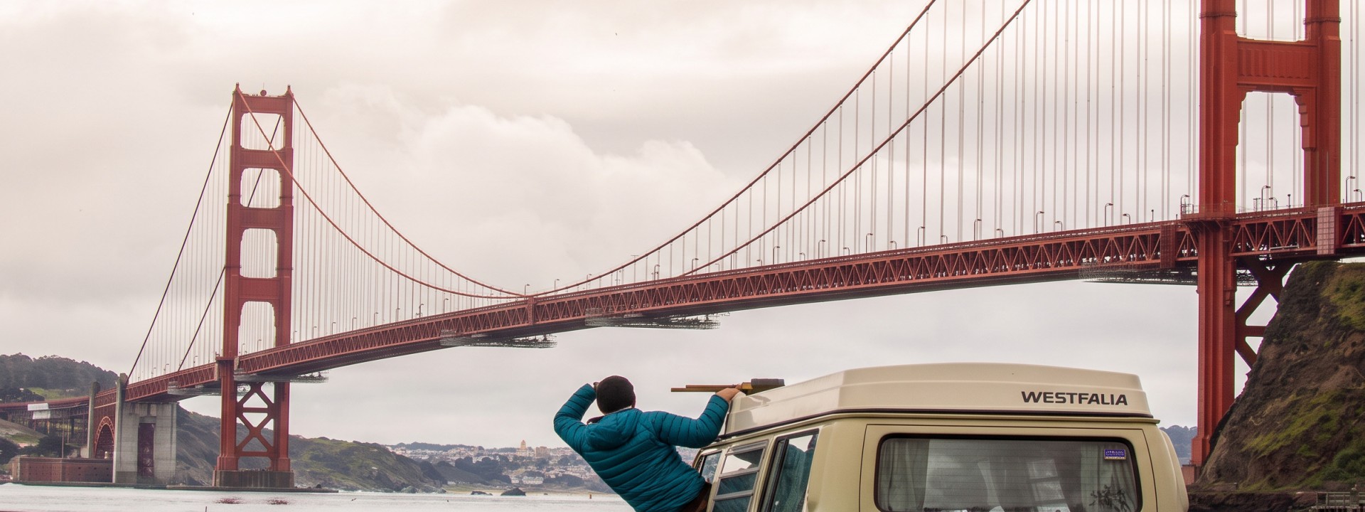 Cloudy sky, the Golden Gate Bridge in San Francisco in the background. A grey-green van is parked on the riverbank, a student in a blue jacket leans out of the window (driver's seat) and looks at the bridge. 