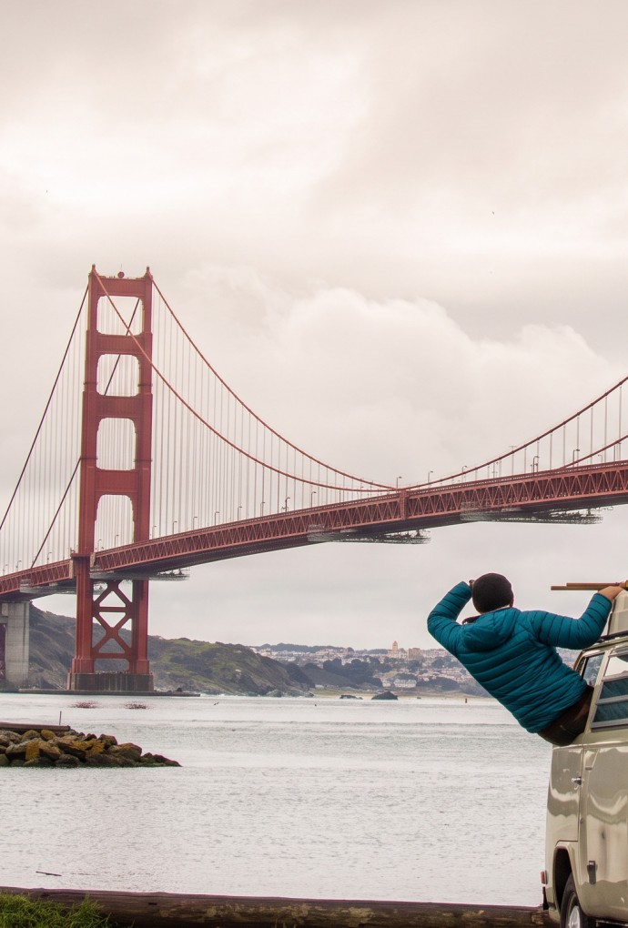 Cloudy sky, the Golden Gate Bridge in San Francisco in the background. A grey-green van is parked on the riverbank, a student in a blue jacket leans out of the window (driver's seat) and looks at the bridge. 