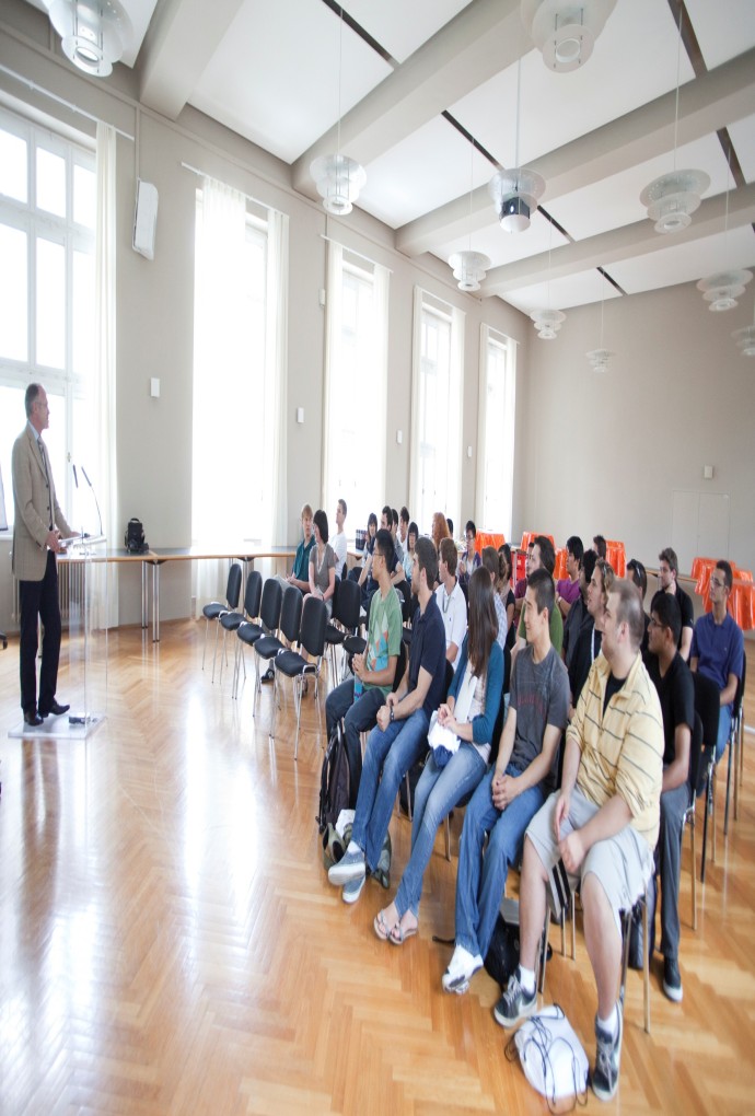 Summer School students sat down on chairs in the Oskar von Miller Room at the Hochschule München, At the front there is a man standing and presenting 