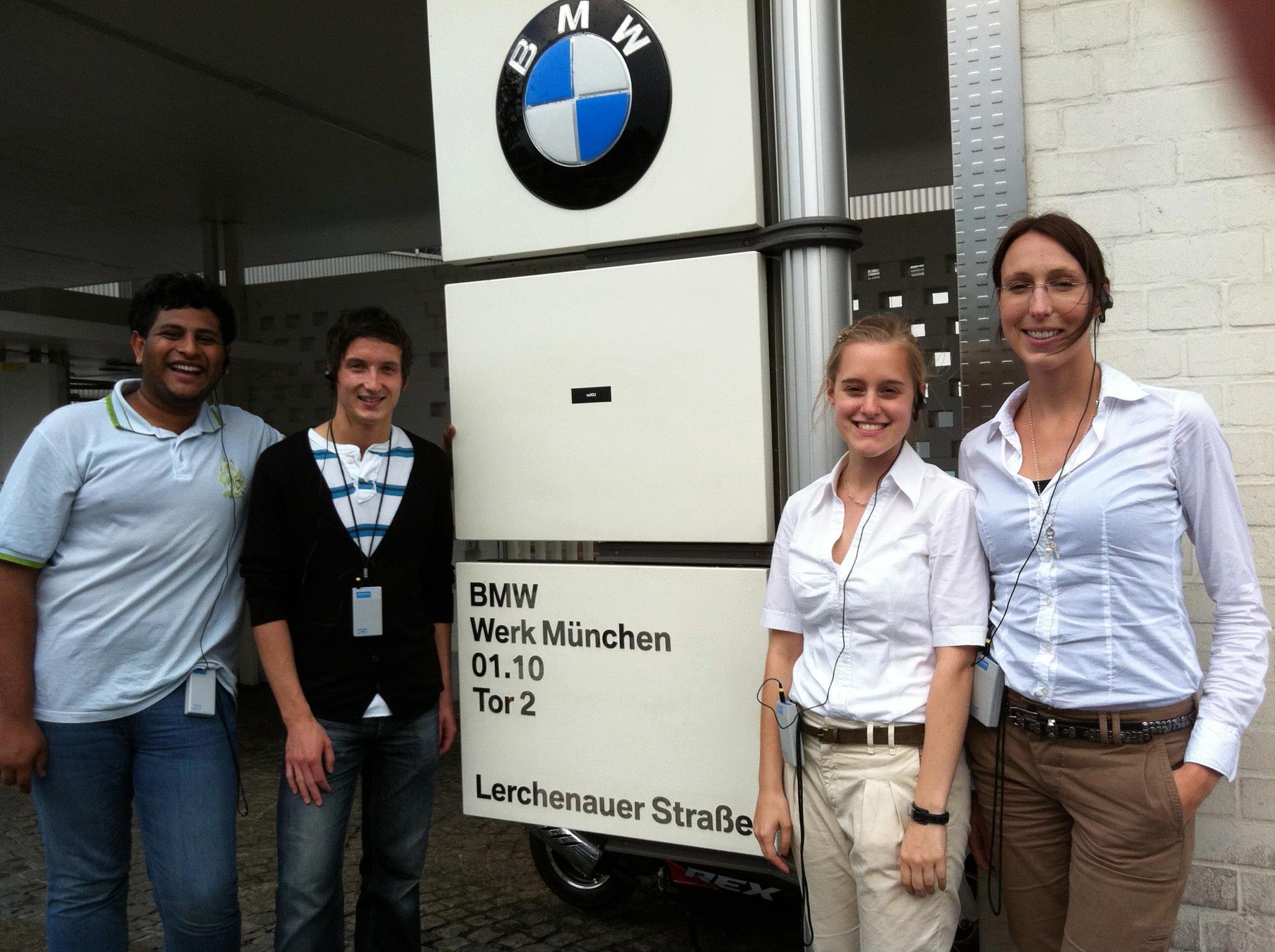 Four smiling students are standing in front of the BMW plant sign with headphones in their ears
