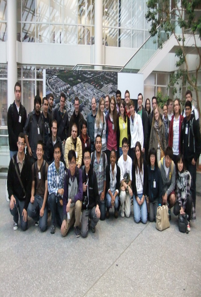 Smiling students stand in front of a picture of the BMW plant, with a tree on the right