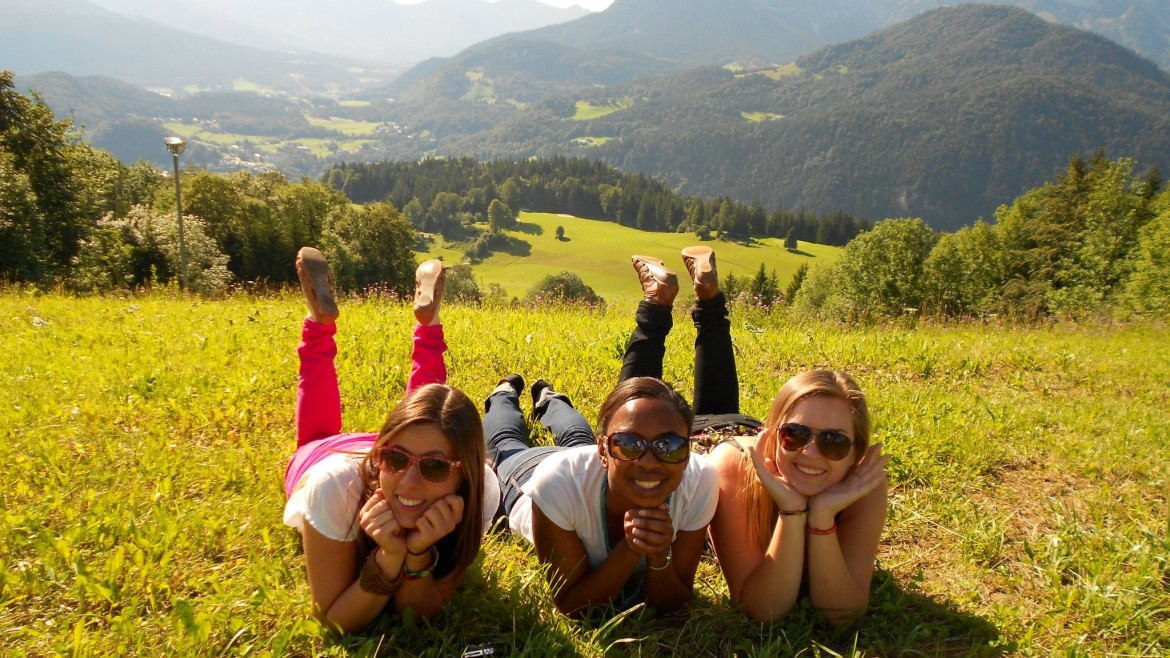 Three girls are lying on a green meadow on a sunny day, with the green mountain scenery in the background