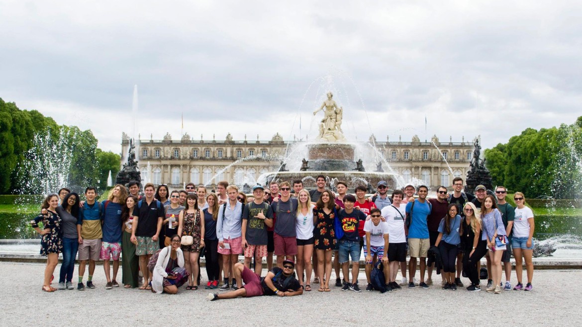 Summer School students posing in front of Herrenchiemsee Palace on a sunny day, with a fountain and trees in the background.