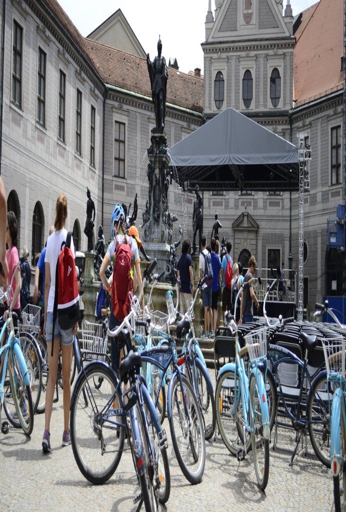 Summer school students standing in front of a fountain with their bikes on a sunny day during the Munich Bike Tour, other people can be seen in the background
