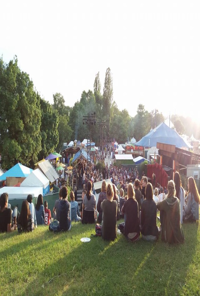 Summer school students sitting on the grass at the Tollwood Festival, with tents and food trucks in the background, on a sunny day