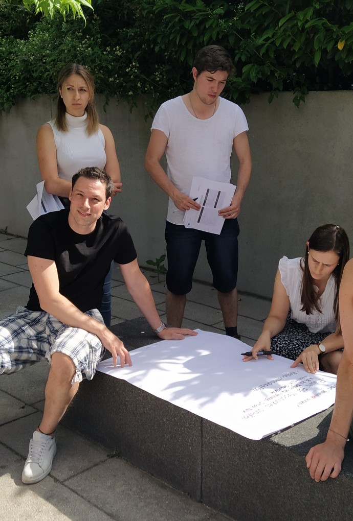 Ein Studierenden-Team sitzt mit einem Flipchart-Papier vor dem T-Gebäude. 