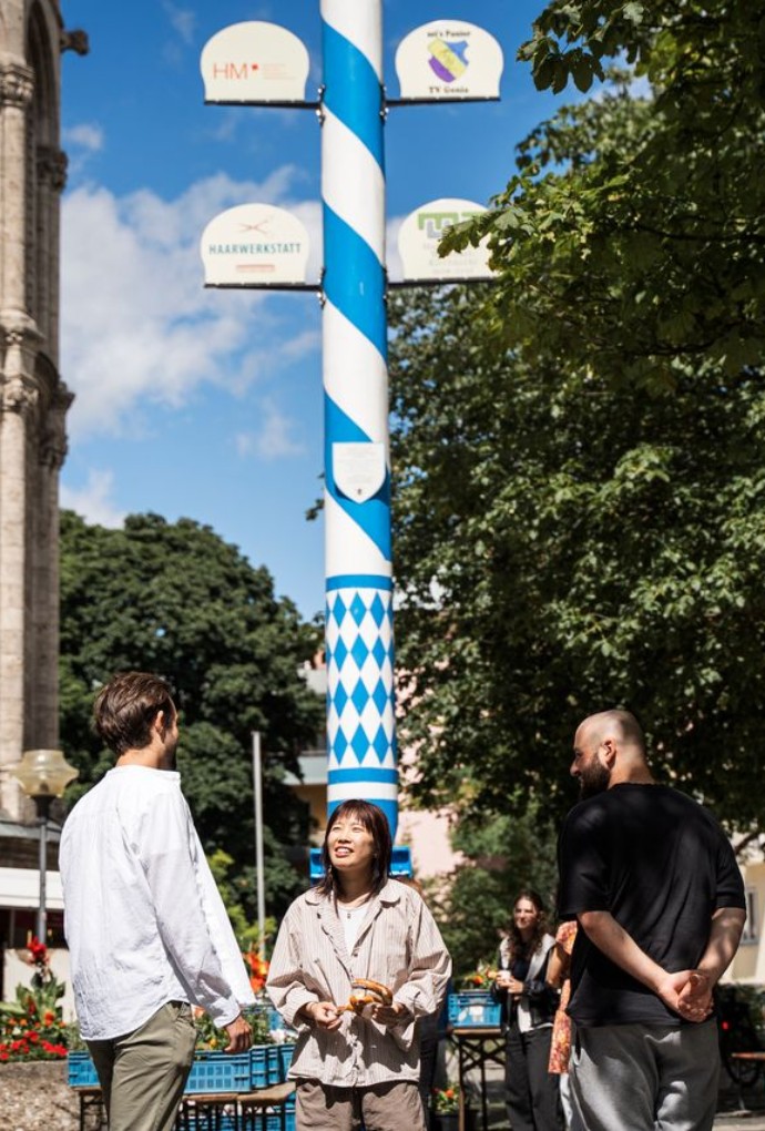 Studierende der Hochschule München stehen auf einem Marktplatz vor dem Maibaum