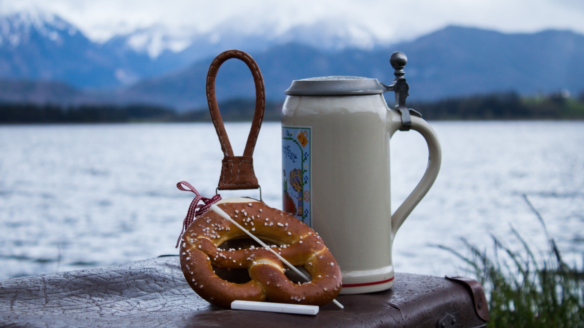 Ein Bierkrug und eine Breze sind vor einem Hintergrund aus See und Bergen.