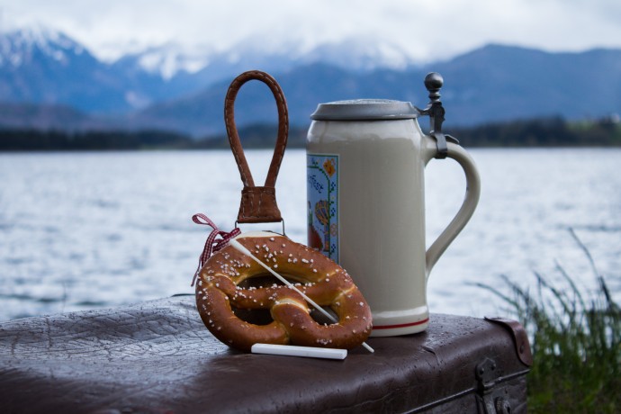 Ein Bierkrug und eine Breze sind vor einem Hintergrund aus See und Bergen.
