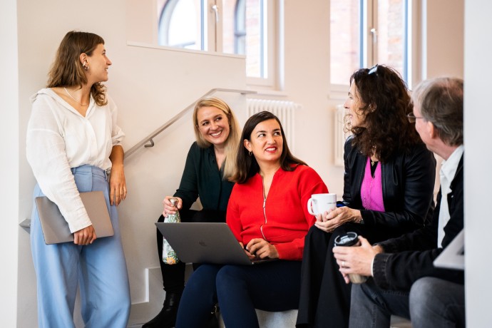 Fünf Mitarbeitende der Hochschule München sitzen auf einer Treppe und lächeln sich zu