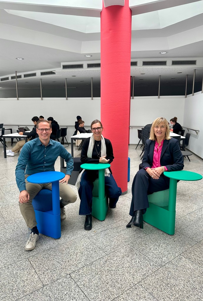 HM-Kanzler Jörg Finger, Prof. Dr. Ruth Maria Otto und HM-Präsidentin Prof. Dr. Sonja Munz sitzen bei der Eröffnung des renovierten Lernzentrums auf neuen, bunten Drehstühlen mit integrierten Tischen.