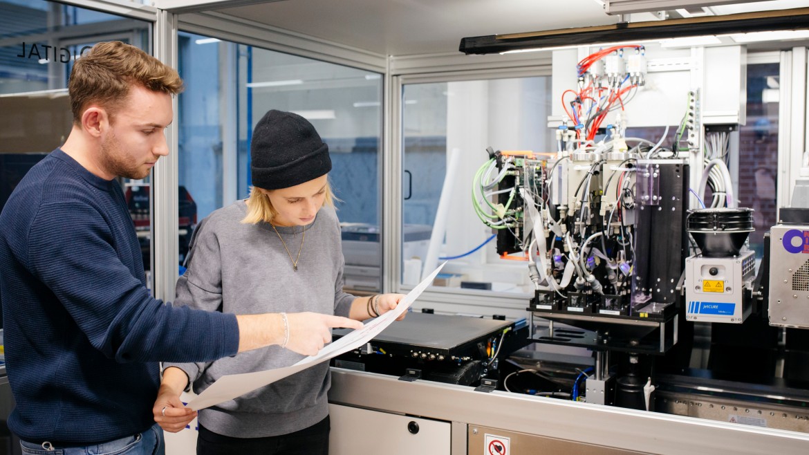Zwei Promovierende stehen vor einer technischen Anlage und betrachten einen Plan.
