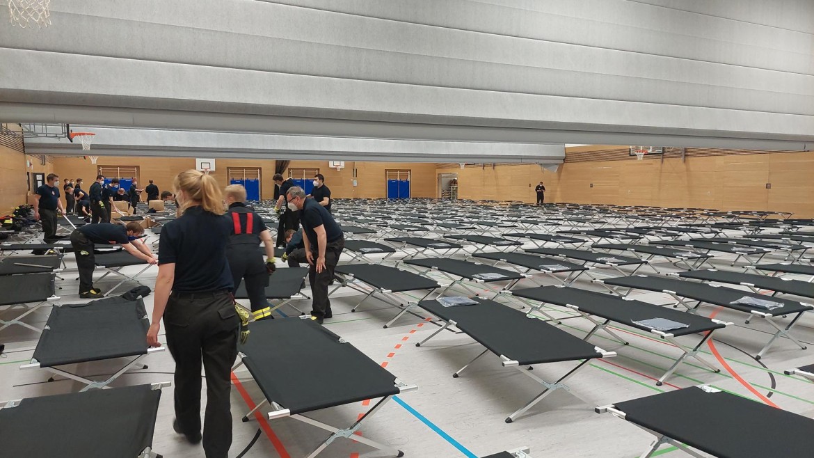 Foto Turnhalle mit aufgestellten Betten und Helferinnen und Helfern