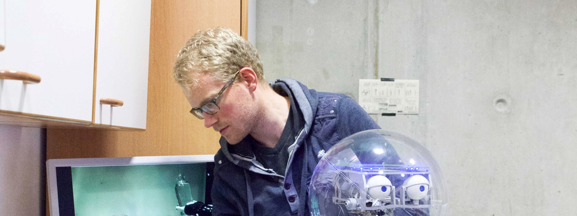 Ein Forscher mit Brille und blauer Kapuzenjacke führt in einer Küchenzeile manuelle Anpassungen an einem Roboterarm durch, welcher eine transparente Plastikflasche greift; der Roboter verfügt über einen transparenten, kugelförmigen Kopf mit sichtbaren Kameramodulen als Augen und ist auf einer fahrbaren Metallsäule montiert, während im Hintergrund ein Spülbecken sowie ein Computermonitor mit einer Live-Übertragung des Greifvorgangs zu sehen sind.