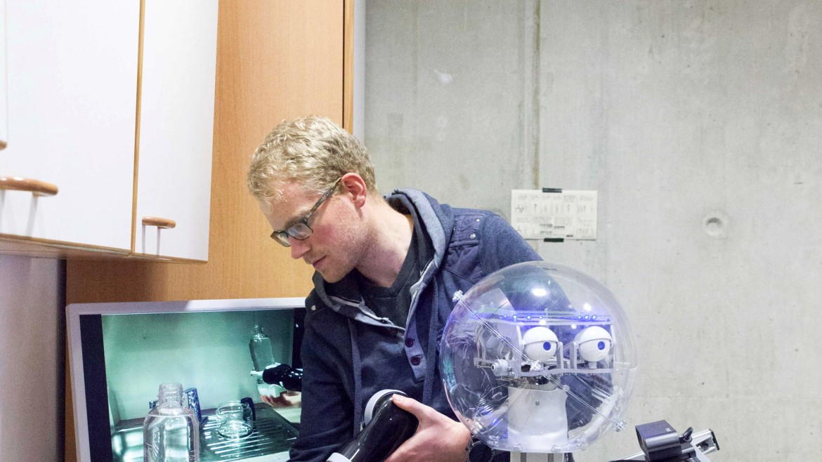 Ein Forscher mit Brille und blauer Kapuzenjacke führt in einer Küchenzeile manuelle Anpassungen an einem Roboterarm durch, welcher eine transparente Plastikflasche greift; der Roboter verfügt über einen transparenten, kugelförmigen Kopf mit sichtbaren Kameramodulen als Augen und ist auf einer fahrbaren Metallsäule montiert, während im Hintergrund ein Spülbecken sowie ein Computermonitor mit einer Live-Übertragung des Greifvorgangs zu sehen sind.