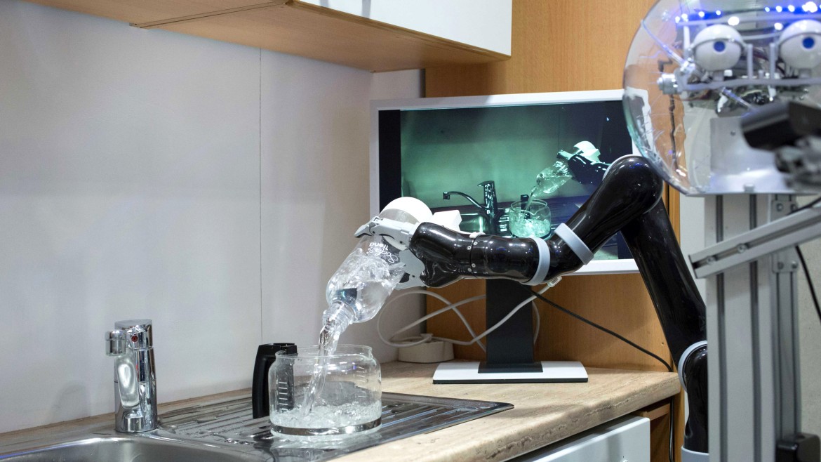 Ein schwarzer Roboterarm mit einer weißen, mehrfingrigen Greifhand gießt im Vordergrund präzise Wasser aus einer transparenten Flasche in eine gläserne Kaffeekanne, die auf der Edelstahlspüle einer Küchenzeile steht; am rechten Bildrand ist der obere Teil des Roboters mit einem transparenten, kugelförmigen Kopf und künstlichen Augen zu erkennen; im Hintergrund liefert ein Computermonitor eine visuelle Rückmeldung des Vorgangs unterhalb von hölzernen Oberschränken.