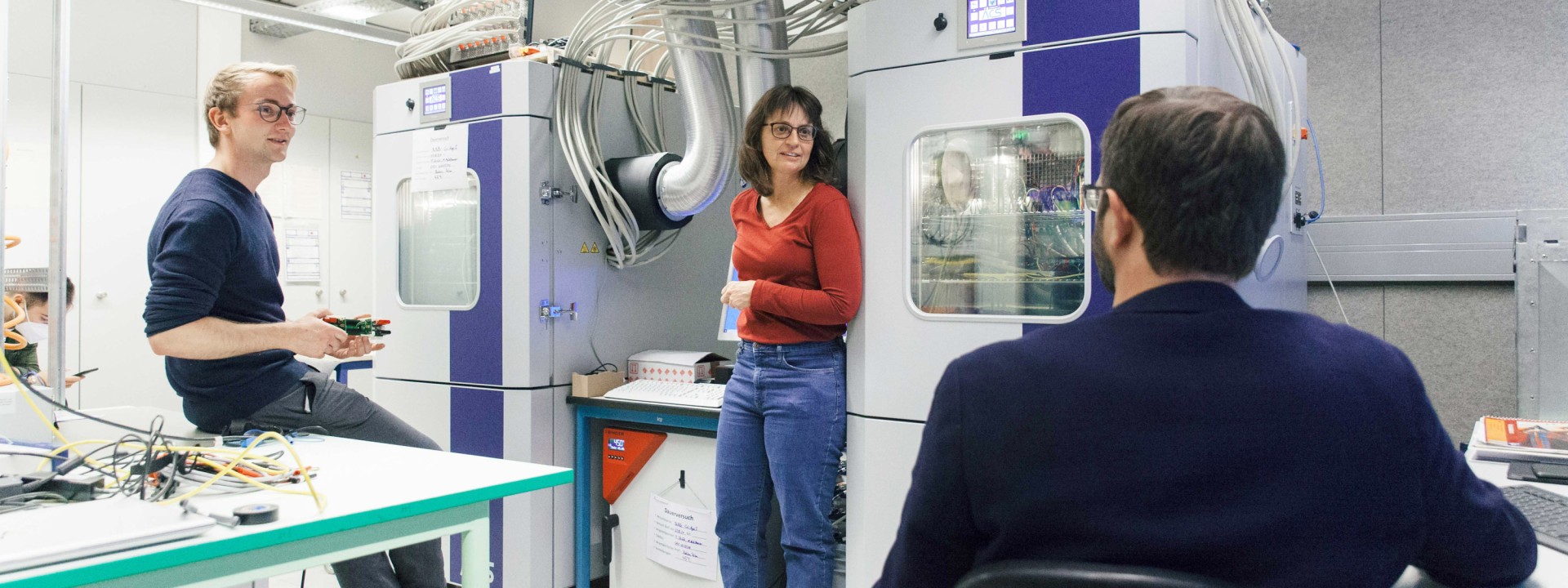 Ein Forscher und eine Forscherin im Gespräch vor zwei ACS-Klimaschrank-Prüfständen in einem Labor. Im Hintergrund sind dicke Alu-Abluftrohre, umfangreiche Kabelbäume und ein Steuerungsmonitor zu sehen.