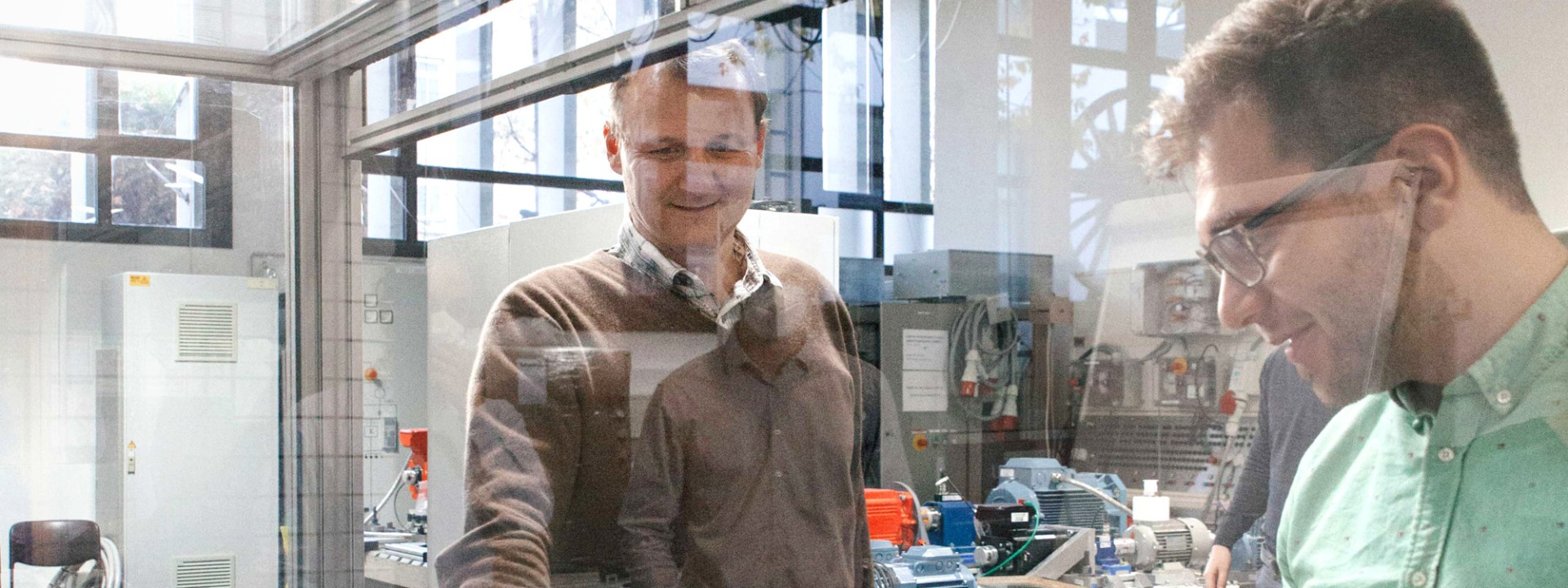 Zwei Forscher in einem Labor für Antriebstechnik oder Leistungselektronik arbeiten an einem Prüfstand hinter einer Sicherheitsglasscheibe, auf dem verkabelte Messgeräte, Leistungskomponenten und Transformatoren angeordnet sind.