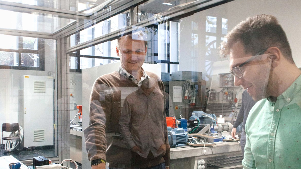 Zwei Forscher in einem Labor für Antriebstechnik oder Leistungselektronik arbeiten an einem Prüfstand hinter einer Sicherheitsglasscheibe, auf dem verkabelte Messgeräte, Leistungskomponenten und Transformatoren angeordnet sind.