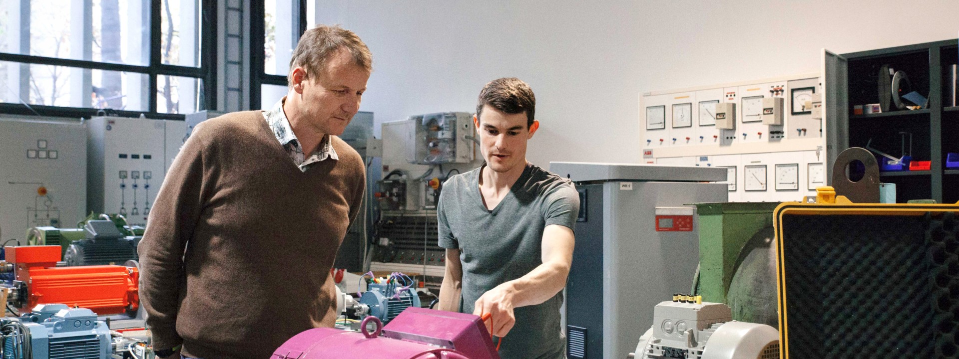 Ein Forscher und ein Student arbeiten im Maschinenlabor an einem Prüfstand mit einer großen, violetten Elektromaschine, während im Hintergrund Schaltschränke, weitere Elektromotoren und ein Laptop zur Messdatenerfassung sichtbar sind.