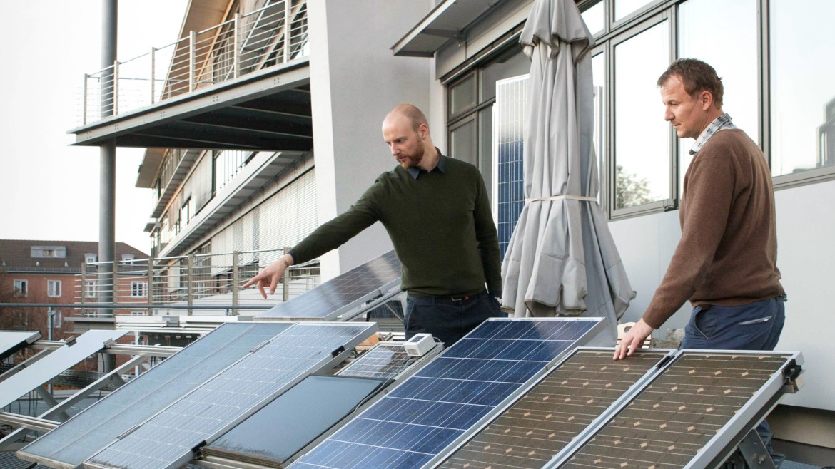 Zwei Forscher überprüfen eine Testanlage mit verschiedenen Photovoltaik-Modulen auf einer Dachterrasse; im Vordergrund steht ein geöffneter, gelber Messkoffer mit angeschlossener Verkabelung.