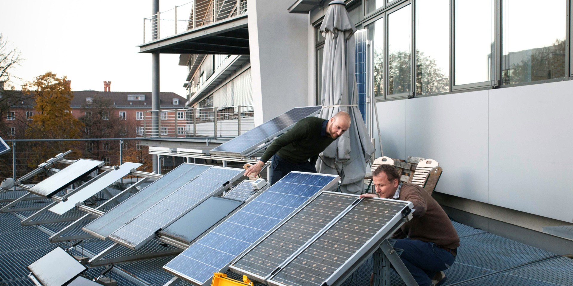 Zwei Forscher inspizieren aus nächster Nähe eine Testreihe verschiedener Photovoltaik-Module auf einer Dachterrasse, während im Vordergrund ein gelber, geöffneter Messkoffer mit Verkabelung positioniert ist.