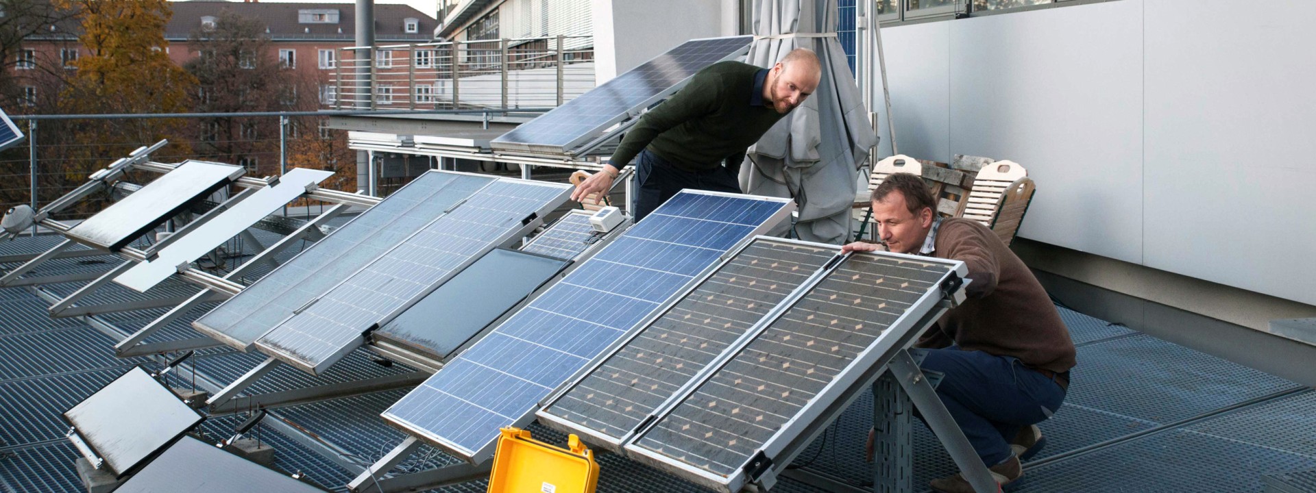 Zwei Forscher inspizieren aus nächster Nähe eine Testreihe verschiedener Photovoltaik-Module auf einer Dachterrasse, während im Vordergrund ein gelber, geöffneter Messkoffer mit Verkabelung positioniert ist.
