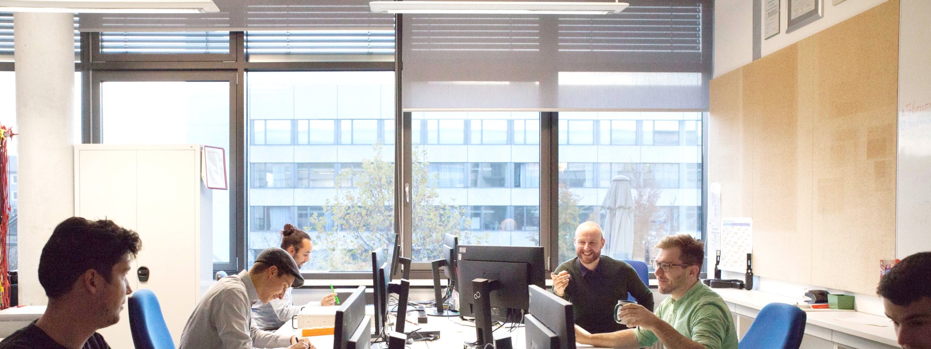 Ein Großraumbüro oder wissenschaftlicher Arbeitsraum mit mehreren Forschern an einer langen Schreibtischreihe, die an PCs arbeiten oder sich im Hintergrund miteinander unterhalten.