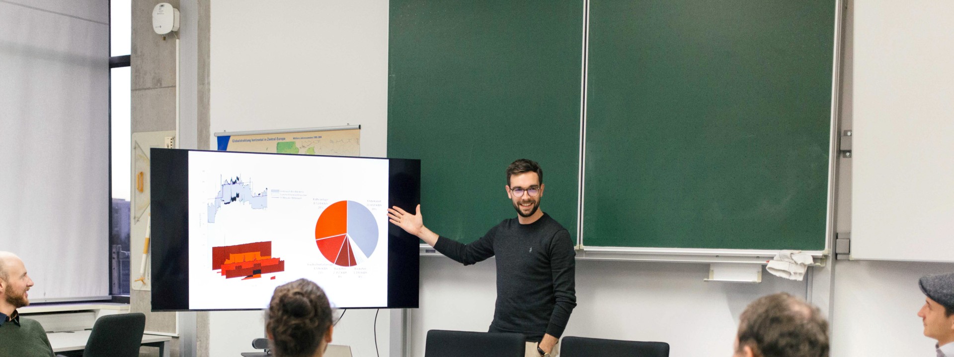 In einem modern ausgestatteten Seminarraum präsentiert ein stehender Forscher mit Brille und schwarzem Pullover, die rechte Hand einladend zu einem großen Monitor mit farbigen Diagrammen und einem Tortendiagramm ausgestreckt, vor einer Runde von fünf sitzenden Kollegen an einem Konferenztisch, während im Hintergrund zwei große, ungenutzte grüne Kreidetafeln und eine Landkarte an der Wand sichtbar sind.