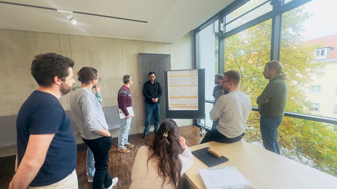 In einem von Tageslicht durchfluteten Seminarraum mit Sichtbetonwand und einer großen Fensterfront steht eine Gruppe von Forschenden um eine Stellwand herum; ein Referent im schwarzen Hemd steht im Hintergrund neben dem aufgestellten wissenschaftlichen Forschungsposter und präsentiert die Inhalte, während sechs Teilnehmende – teils stehend, teils am Rand eines hellen Holztisches sitzend – aufmerksam zuhören; im Vordergrund sitzt eine Frau mit langen, braunen Haaren mit dem Rücken zur Kamera am Tisch, auf dem Notizen, ein Kaffeebecher und ein Smartphone liegen, während der Blick durch das Fenster auf herbstliche Bäume fällt.