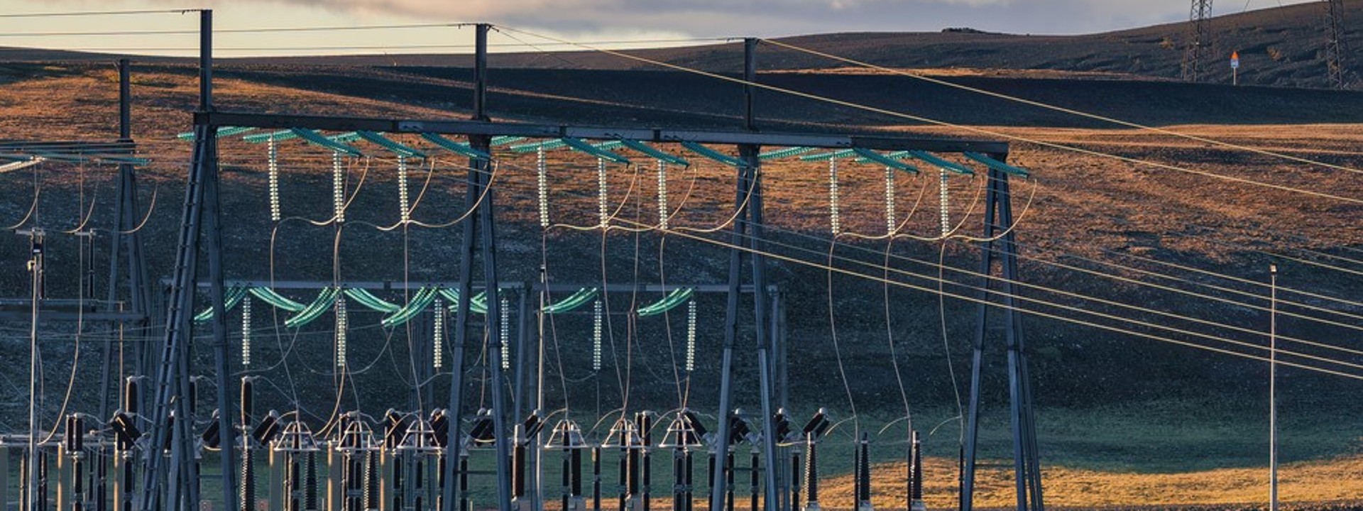Eine weitläufige Freiluftschaltanlage mit metallischen Gerüststrukturen, Isolatorenketten und dichten Leitungsbündeln steht im linken Bildvordergrund einer hügeligen, kargen Naturlandschaft; am rechten Bildrand erstreckt sich ein hoher Gittermast einer Hochspannungsleitung in die Ferne, während eine dichte Wolkenfront vor einem hellen Himmel im Hintergrund die ländliche Szenerie der elektrischen Energieübertragung einrahmt.