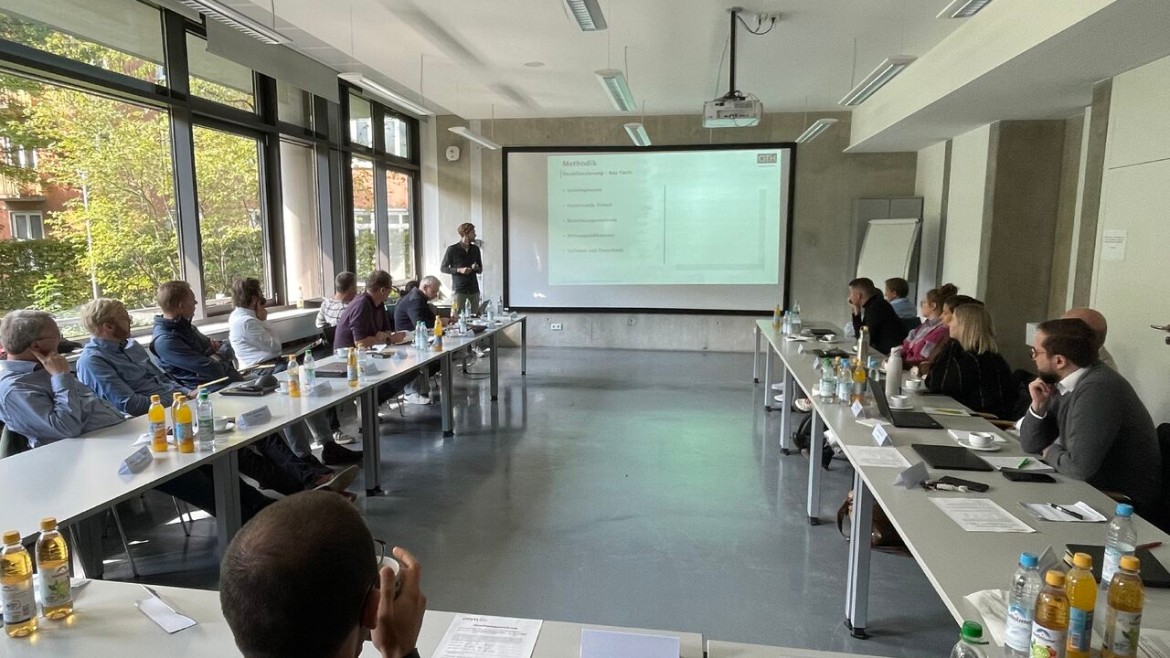 In einem taghellen, modernen Seminarraum mit einer großen Fensterfront auf der linken Seite verfolgt eine Gruppe von sitzenden Forschenden aufmerksam einen Vortrag; die Tische sind in einer U-Form angeordnet, auf denen Getränkeflaschen, Laptops und Notizen verteilt sind; am Kopfende des Raumes steht ein Referent vor einer großen Projektionsleinwand, die eine Präsentationsfolie mit der Überschrift „Methodik“ zeigt, während im Vordergrund der Hinterkopf eines Teilnehmers zu sehen ist.