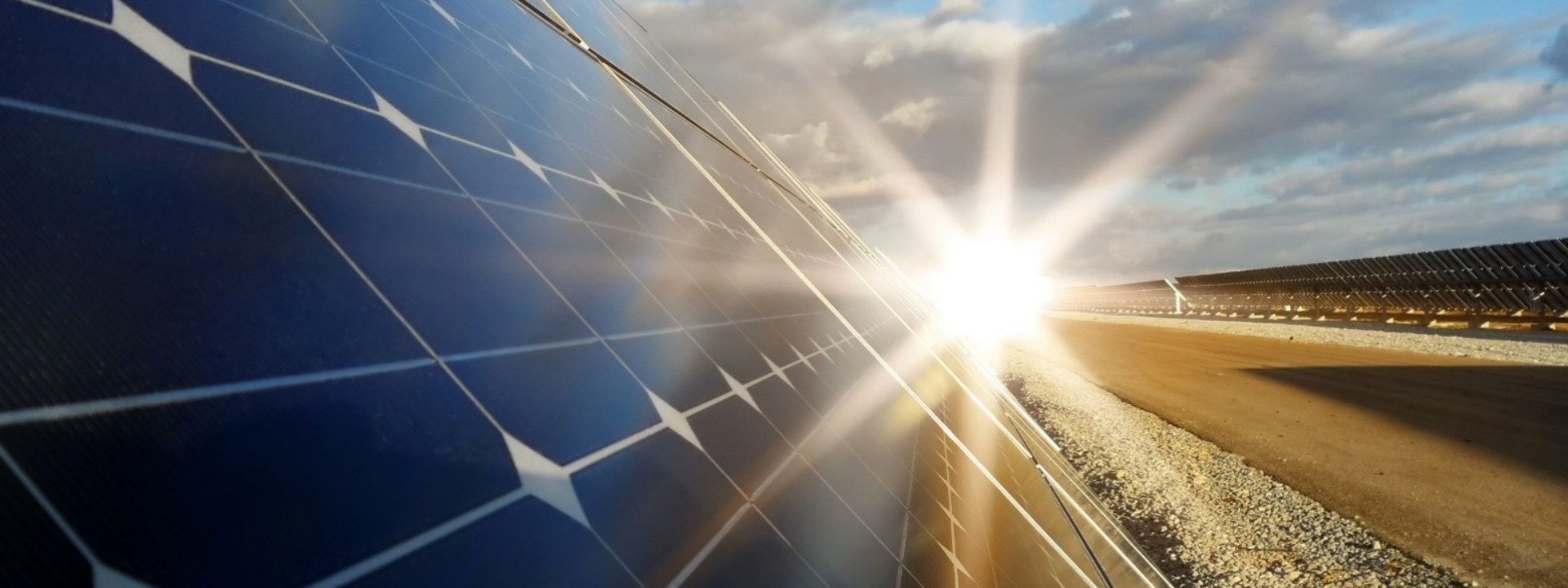 In einer dynamischen, diagonalen Perspektive zeigt die Datei image_363b1b.jpg die Nahaufnahme eines großflächigen Solarmoduls auf einem Freigelände; die tiefblauen, quadratischen Solarzellen mit ihren typischen weißen Eckverbindungen dominieren die linke Bildhälfte, während am rechten Rand des Moduls die Sonne intensiv hervor bricht und sternförmige Lichtstrahlen (Lens Flare) über die gesamte Szenerie wirft; im rechten Hintergrund erstreckt sich ein Schotterweg neben weiteren, flach aufgestellten Modulreihen eines Solarparks unter einem leicht bewölkten, blauen Himmel.