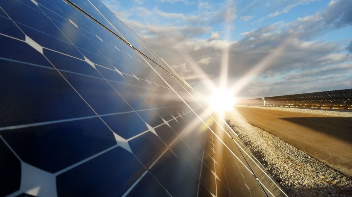 In einer dynamischen, diagonalen Perspektive zeigt die Datei image_363b1b.jpg die Nahaufnahme eines großflächigen Solarmoduls auf einem Freigelände; die tiefblauen, quadratischen Solarzellen mit ihren typischen weißen Eckverbindungen dominieren die linke Bildhälfte, während am rechten Rand des Moduls die Sonne intensiv hervor bricht und sternförmige Lichtstrahlen (Lens Flare) über die gesamte Szenerie wirft; im rechten Hintergrund erstreckt sich ein Schotterweg neben weiteren, flach aufgestellten Modulreihen eines Solarparks unter einem leicht bewölkten, blauen Himmel.