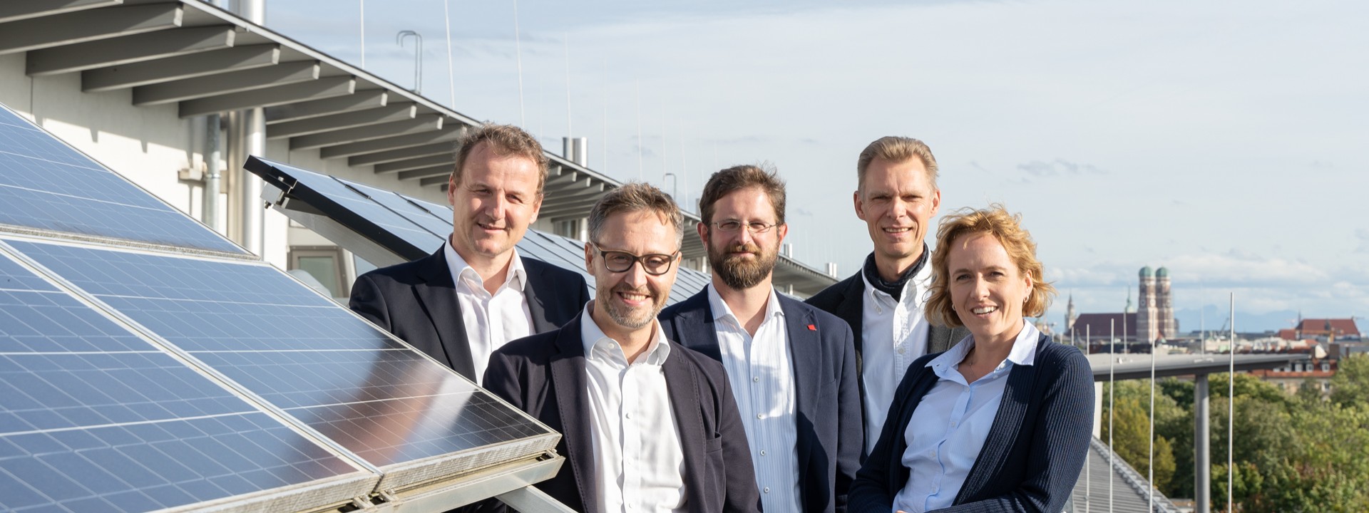 Ein Gruppenfoto zeigt die fünf Gründer des „Institute for Sustainable Energy Systems“ (ISES) auf einer modernen Dachterrasse im Freien; das Team – bestehend aus vier Männern in Sakkos und hellen Hemden sowie einer Frau in einer dunkelblauen Strickjacke und hellblauem Hemd – blickt lächelnd in die Kamera; auf der linken Bildseite ist im Vordergrund eine aufgestellte Photovoltaikanlage montiert, deren glänzende Solarmodule schräg ins Bild ragen; im rechten Hintergrund erstreckt sich hinter Bäumen die Silhouette einer Stadt, bei der die markanten Türme der Münchener Frauenkirche unter einem leicht bewölkten Himmel gut zu erkennen sind.